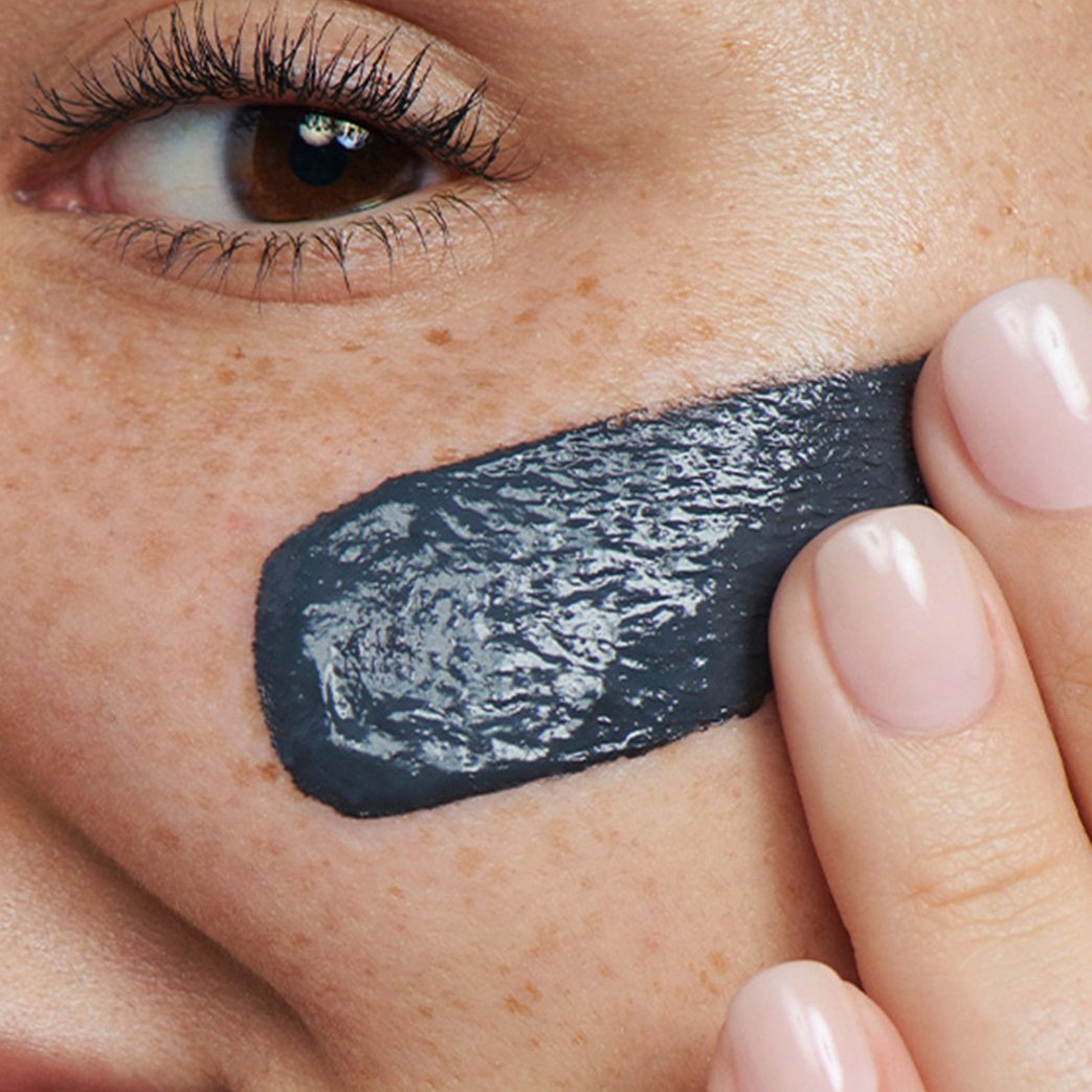 Close-up of a person applying a dark grey charcoal mask to their cheek.